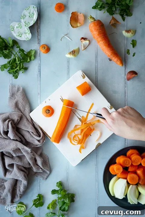 A carrot being chopped on a white chopping board with a pairing knife. There's lots of vegetable peelings and coriander scattered around, a light brown linen napkin, some stock pots and a plate filled with chopped carrot and onion. All set on a cool grey wood effect backdrop.