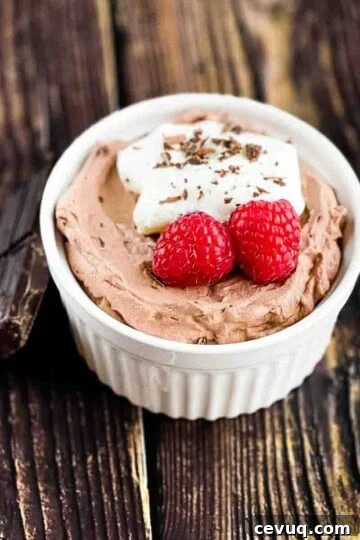 Keto chocolate mousse in a white ramekin, with a dollop of whipped cream, two raspberries, and shaved chocolate for garnish.