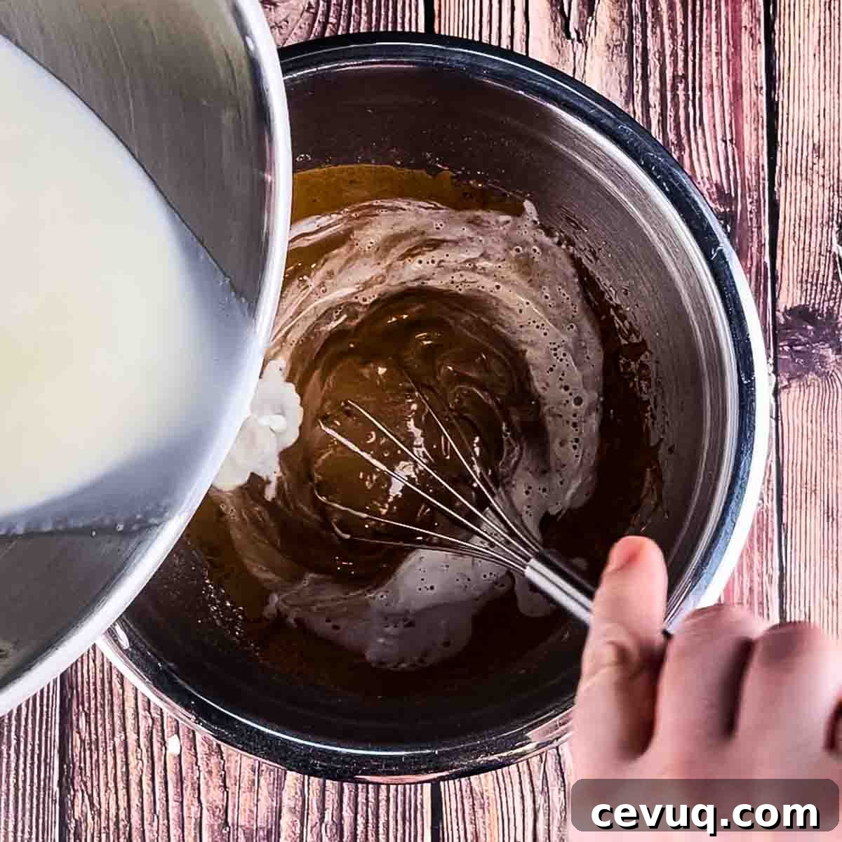 The hot cream being poured into the mixing bowl to temper the eggs, a hand using a whisk to mix.