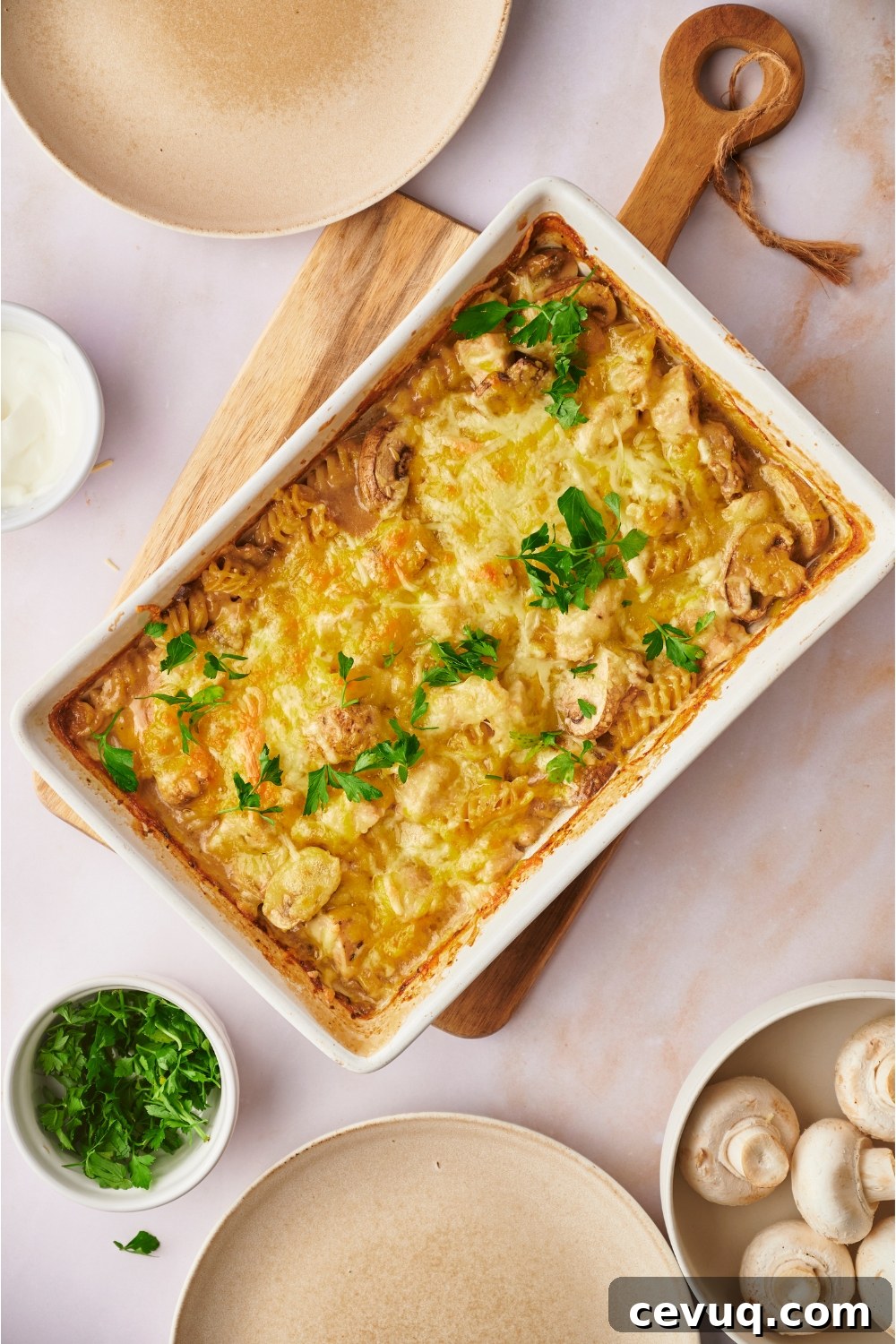 Chicken and mushroom casserole freshly baked in a white baking dish, garnished with fresh herbs, on a wooden board. Two empty plates surround the baking dish.