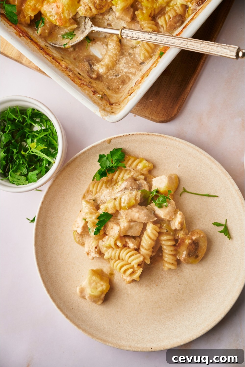 A serving of chicken and mushroom casserole garnished with fresh herbs on an off-white plate. A bowl of fresh herbs and the rest of the casserole surround the plate.