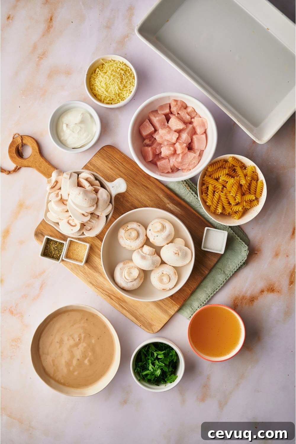An assortment of ingredients including bowls of raw mushrooms, raw chicken, dried pasta, broth, fresh herbs, cheese, and sour cream.