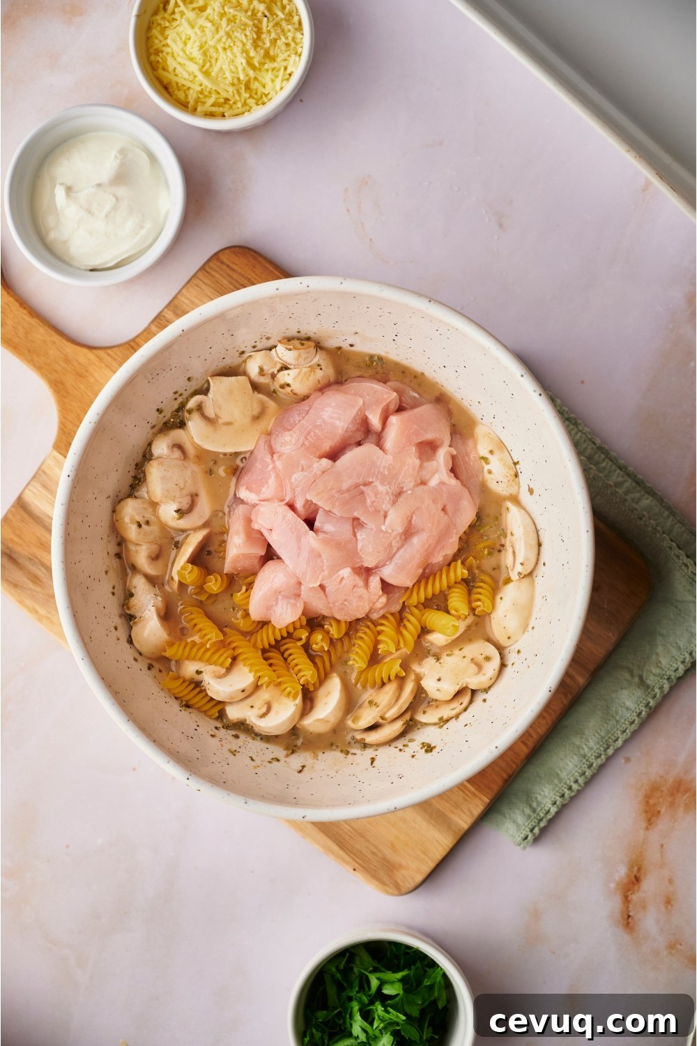Large white bowl filled with raw diced ham, uncooked pasta, sliced mushrooms, herbs, sliced mushrooms, and a creamy soup mixture.