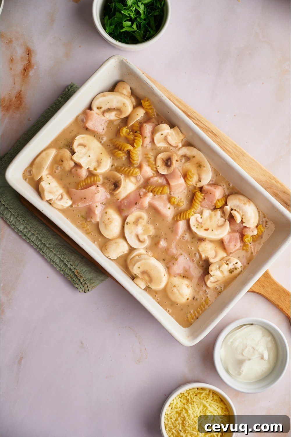 White baking dish on top of a wooden board filled with unbaked chicken and mushroom casserole.