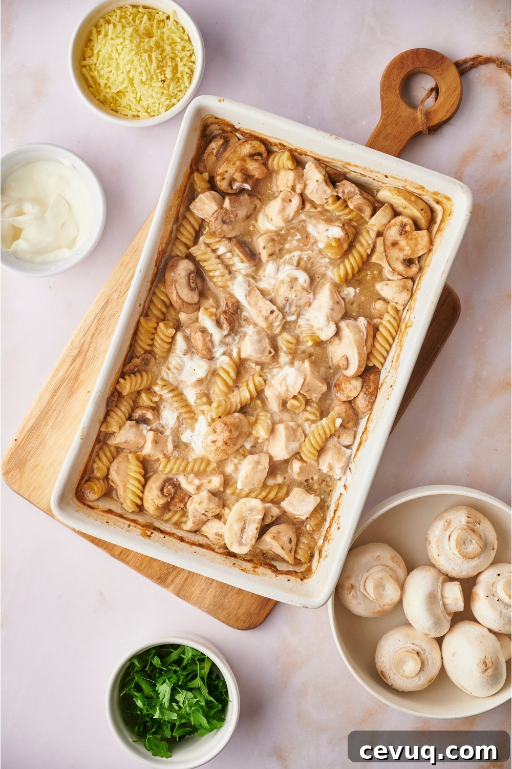 White baking dish on top of a wooden board filled with freshly baked chicken and mushroom casserole that has sour cream mixed on top. The dish is surrounded by ingredients including bowls of mushrooms, sour cream, and herbs.