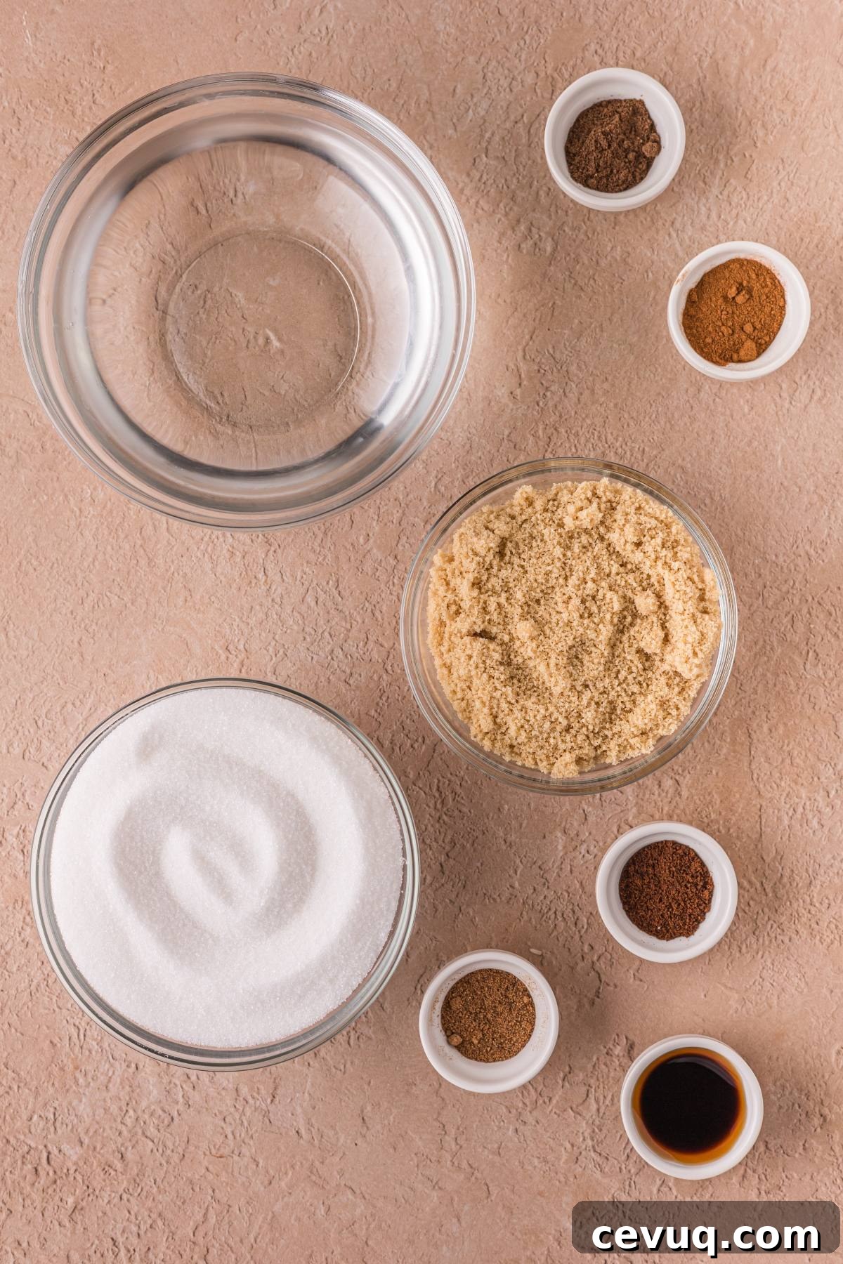 Overhead view of clear bowls with sugar, brown sugar, vanilla, and spices on a tan surface.