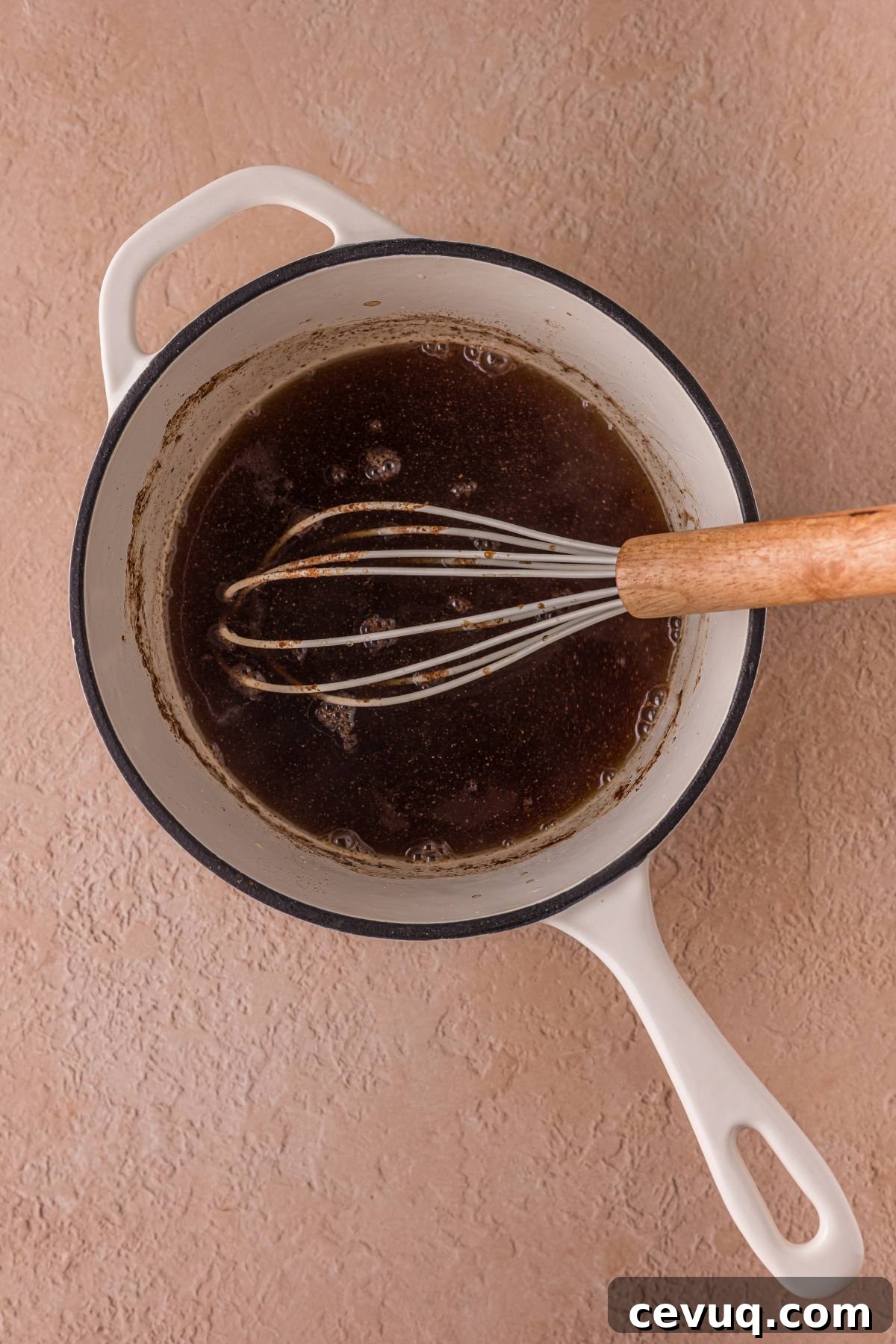A saucepan with a whisk stirring a dark liquid on a light textured surface.