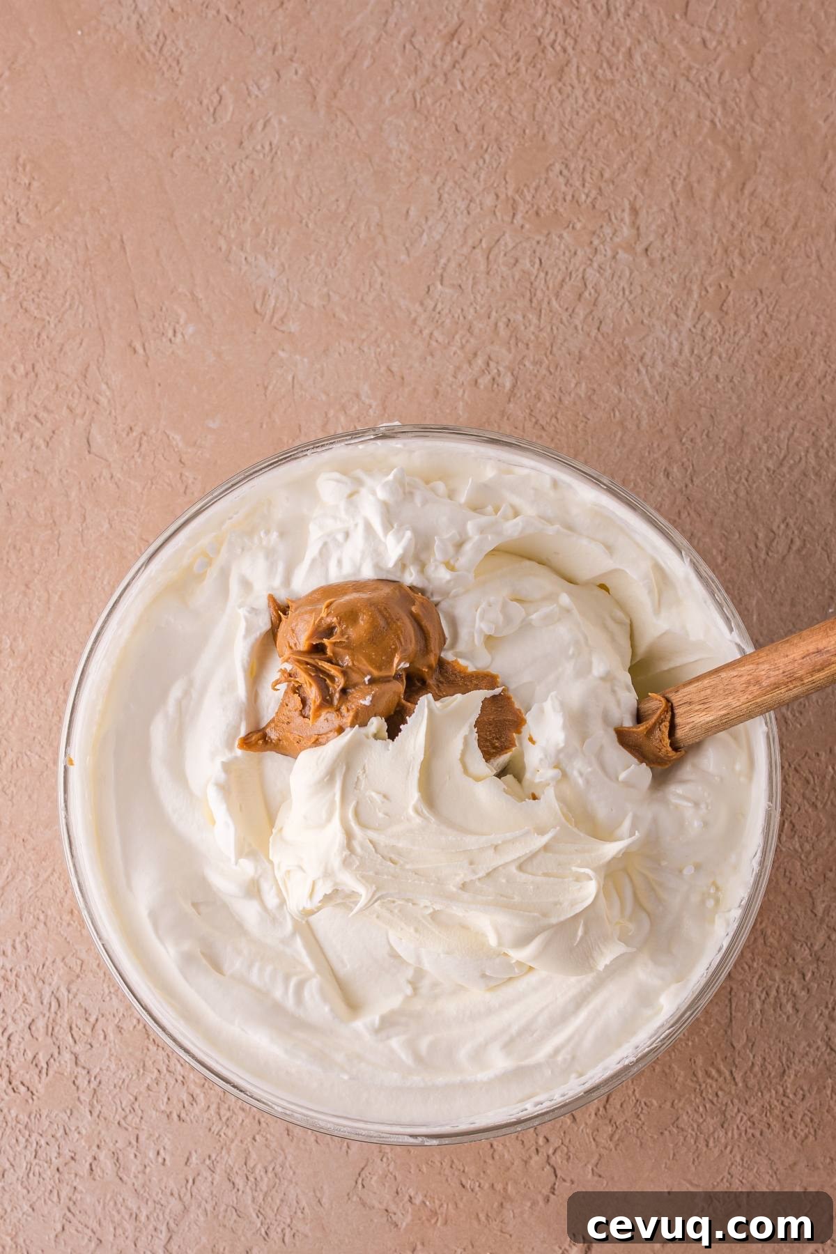 A glass bowl of whipped cream with a dollop of peanut butter and a wooden spoon on a textured surface.