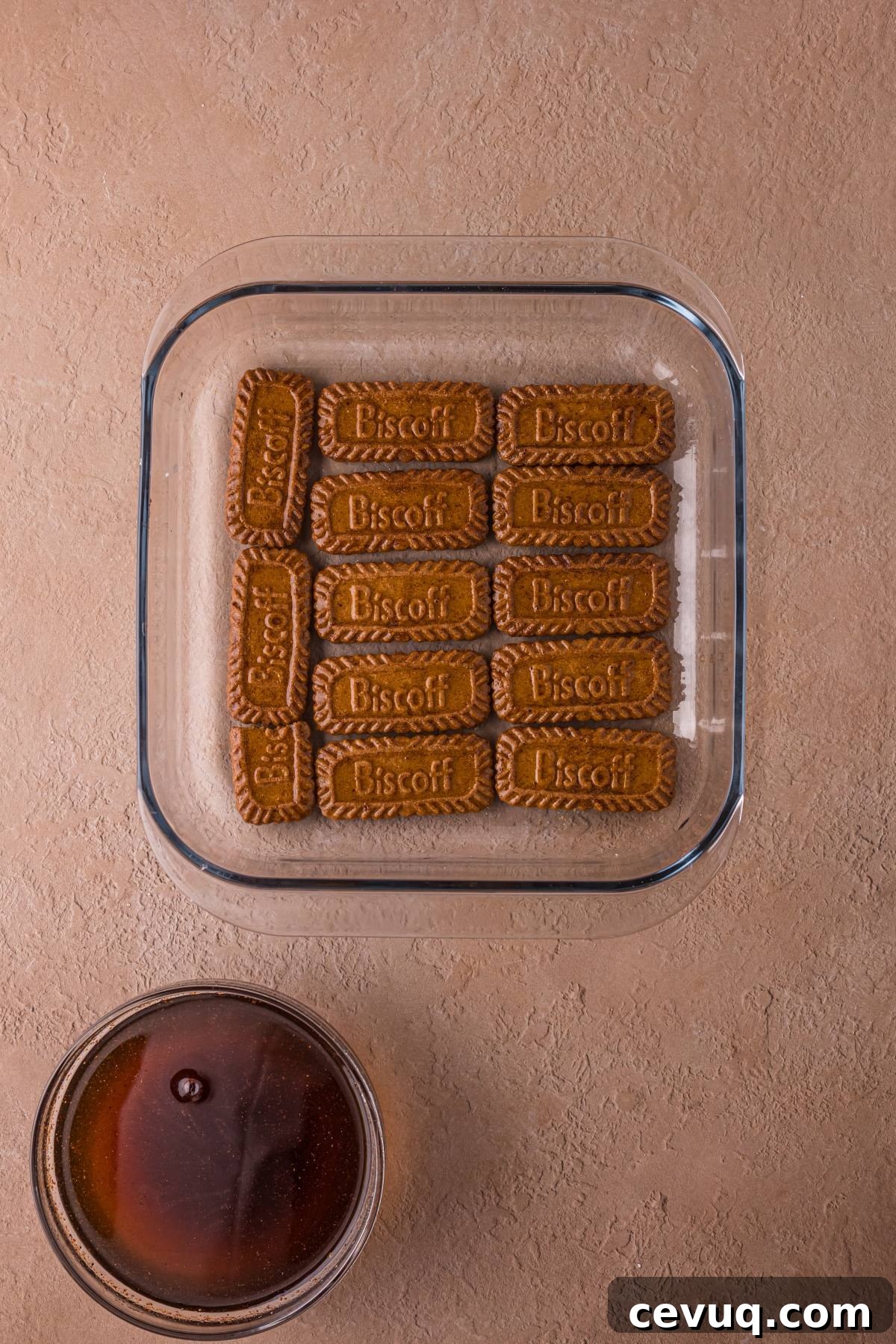 Square glass dish with a layer of Biscoff cookies next to a bowl of dark liquid on a beige surface.