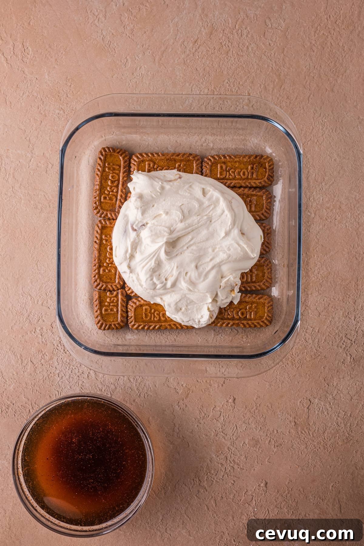 A glass dish with cookies topped by cream, next to a bowl of dark liquid on a beige surface.