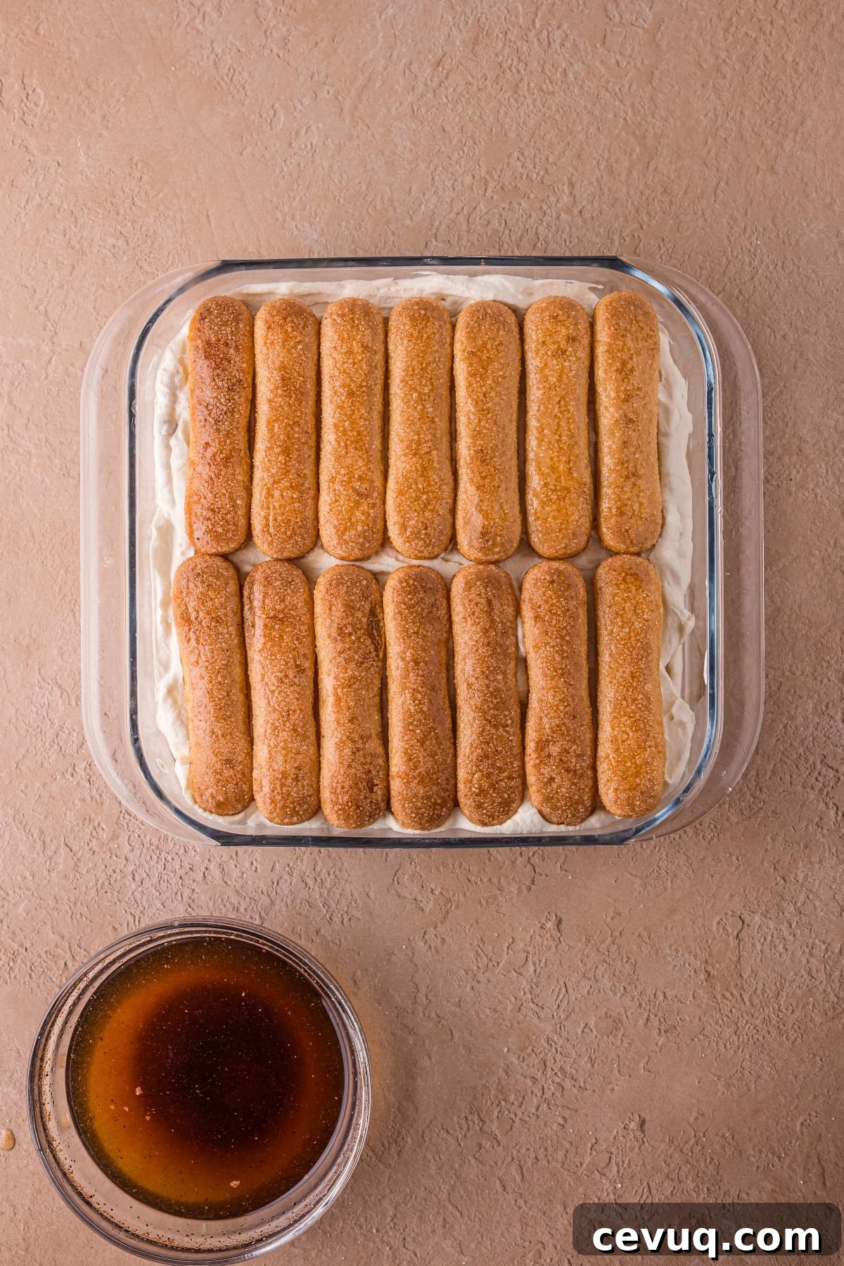A glass dish with a layer of ladyfingers on cream, next to a bowl of coffee on a tan surface.