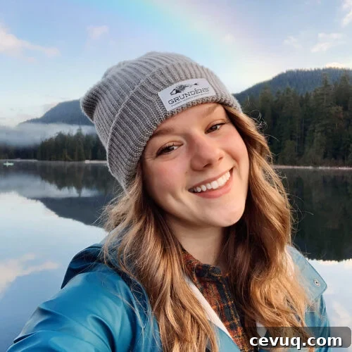 Smiling woman, Bella Bucchiotti, in a gray beanie by a lake with a rainbow and forested mountains in the background.