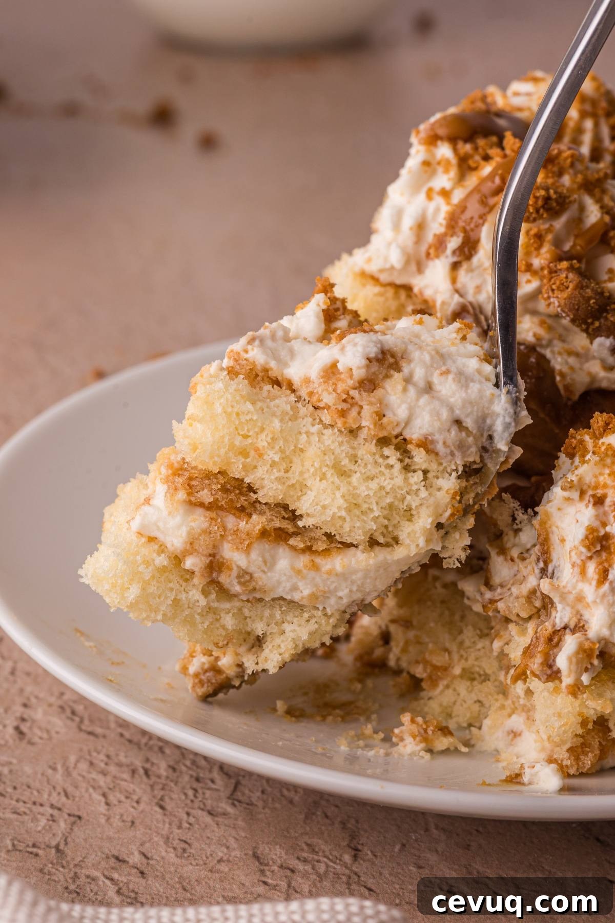 A fork holds a bite of creamy layered cake with whipped topping on a white plate.