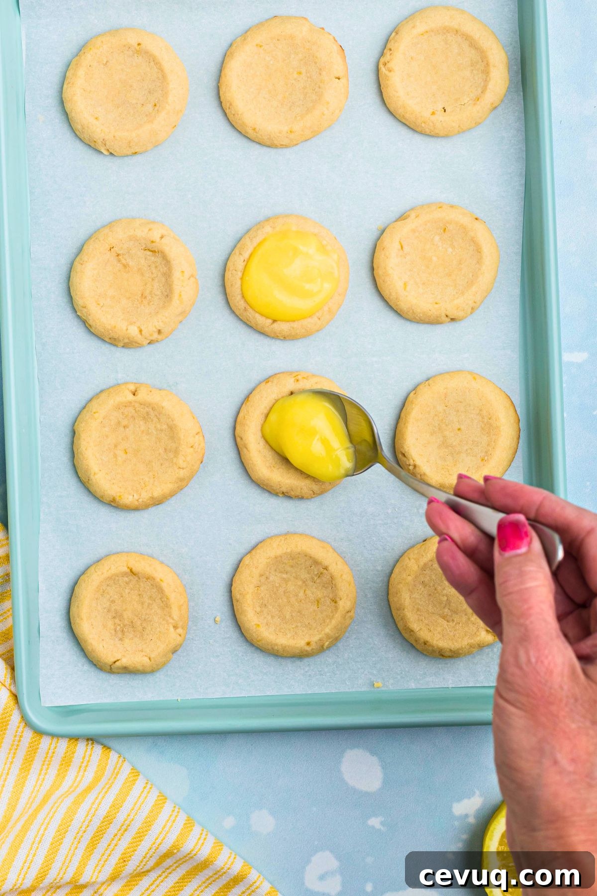 Shortbread cookies being topped with a spoonful of lemon curd, on a baking sheet. 