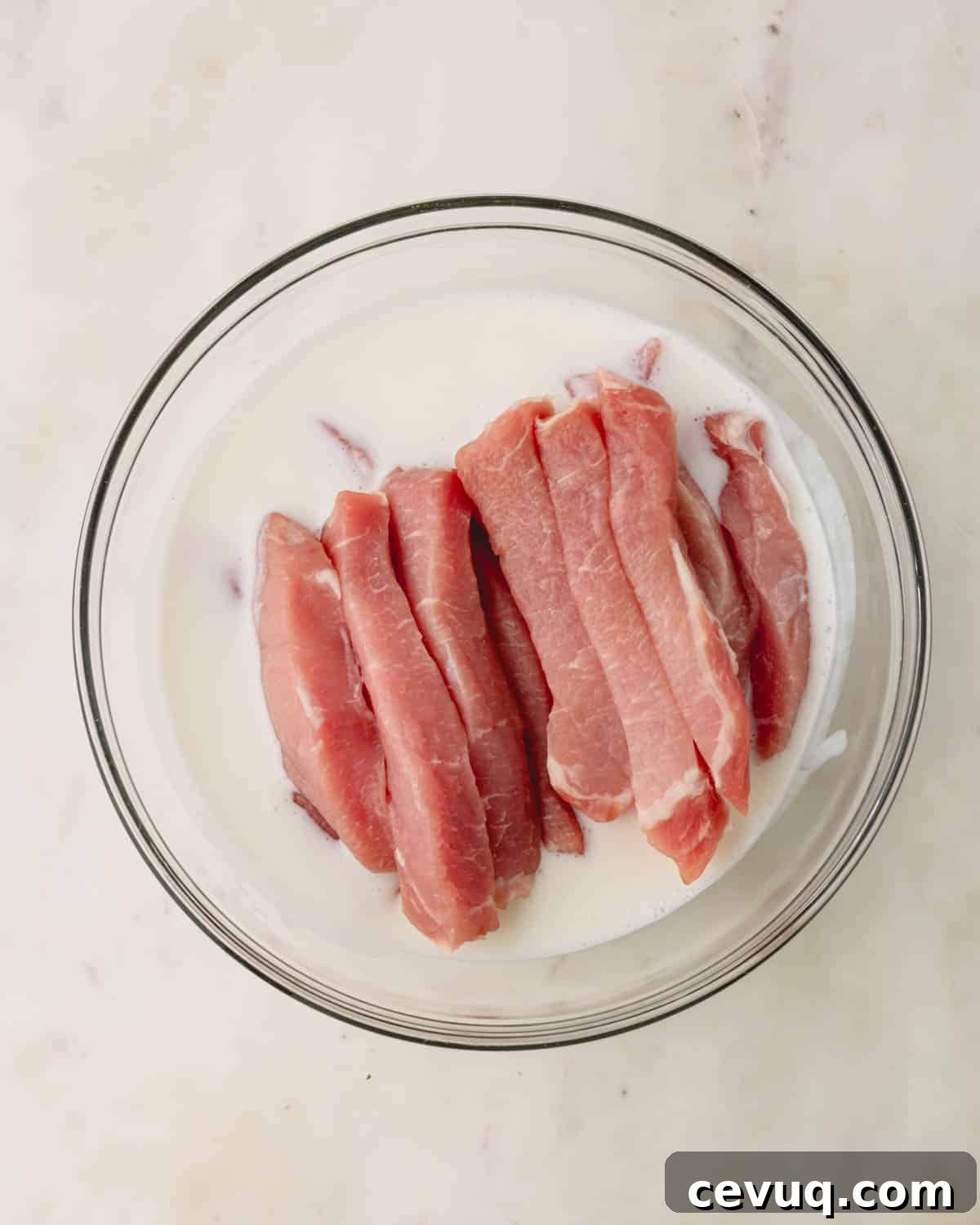 Raw strips of pork tenderloin in a bowl of buttermilk.