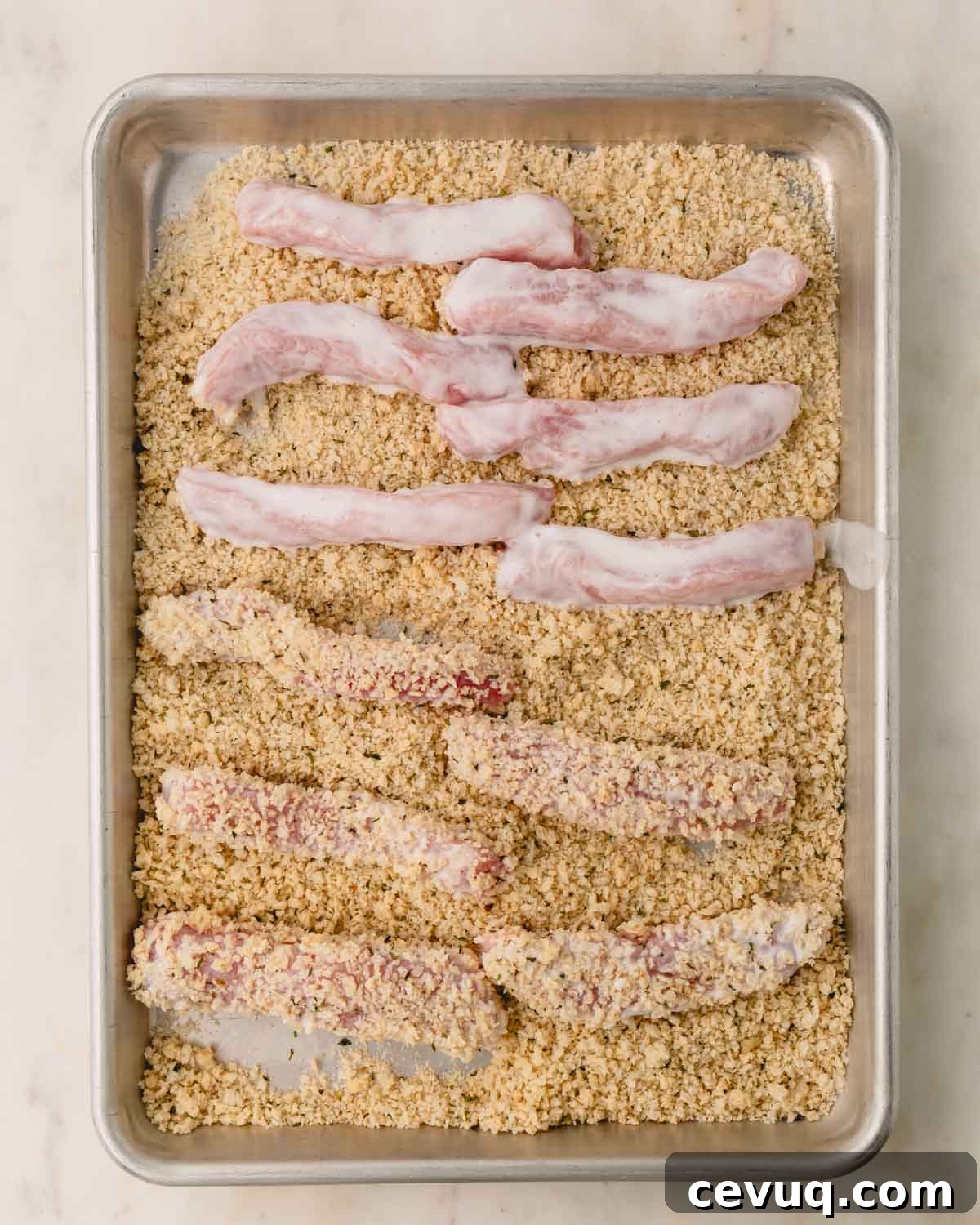 Buttermilk coated pork strips on a baking dish with breadcrumbs.