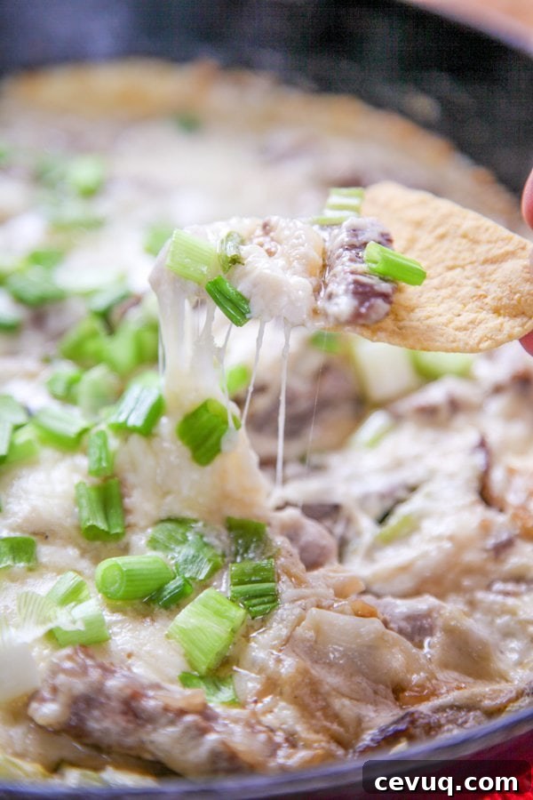 a torilla chip dipping into cheesy steak dip topped with cheese and green onions.