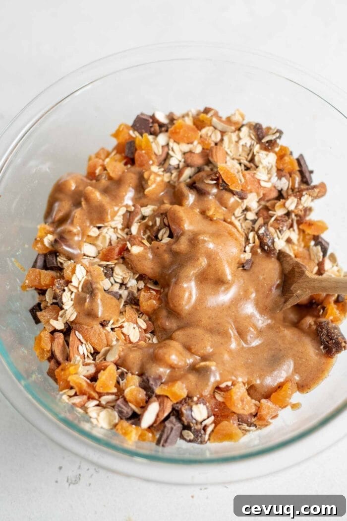 Almonds, oats, dates, apricots and chocolate with maple syrup in a glass mixing bowl.