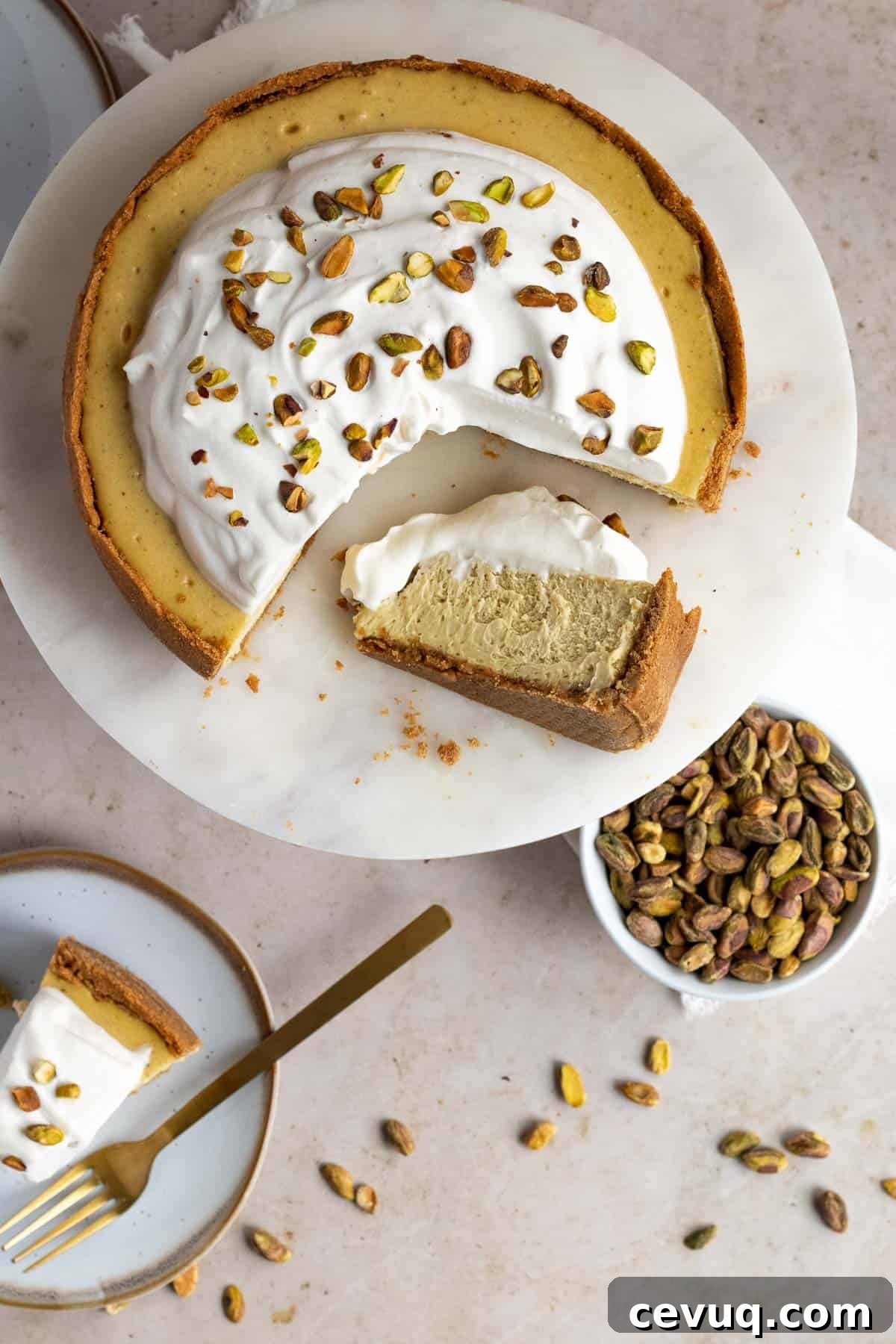 top view of a pistachio cheesecake on a marble cake stand with a slice on a plate and a bowl of pistachios nearby
