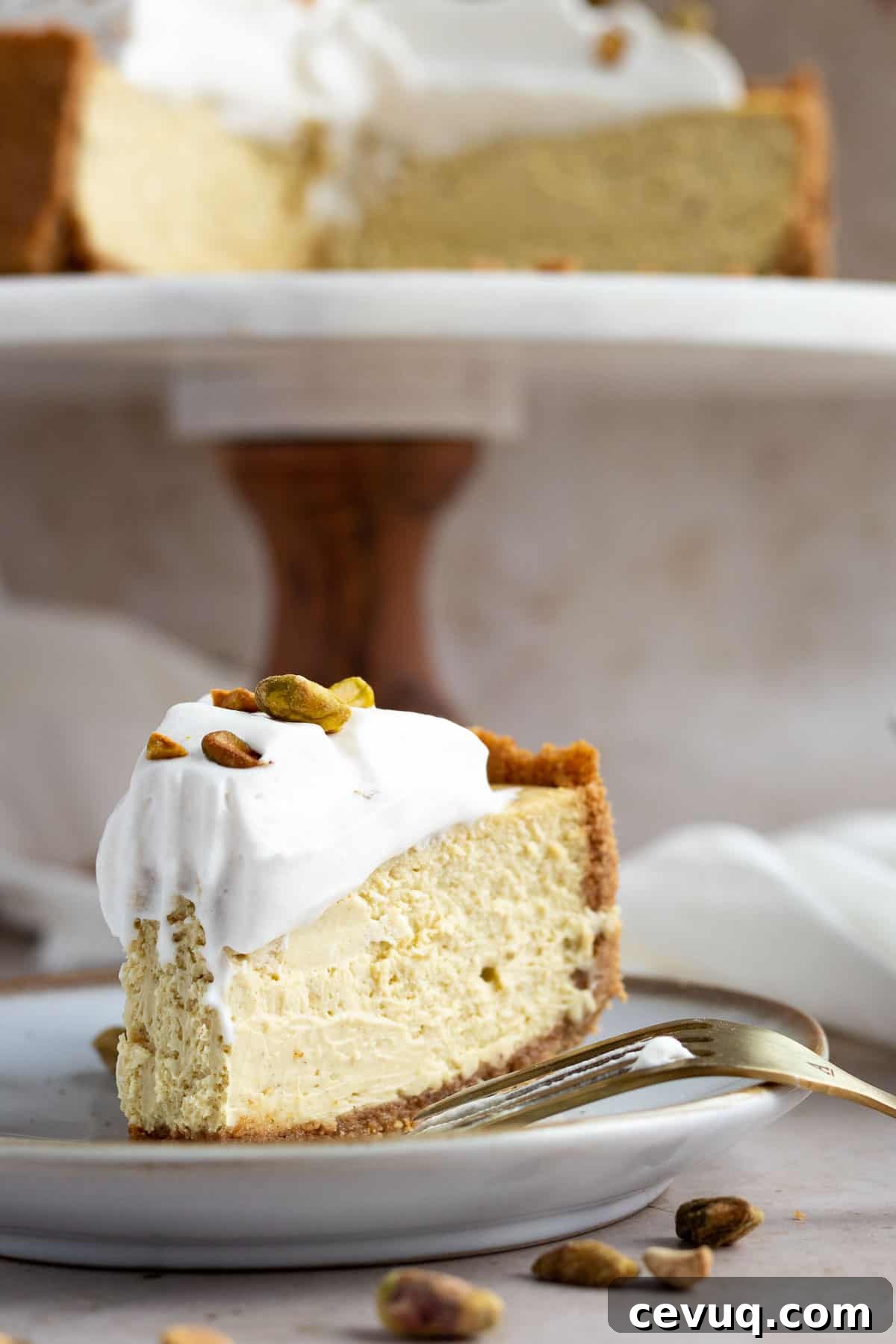 pistachio cheesecake slice on a plate with the whole cheesecake on a cake stand behind it