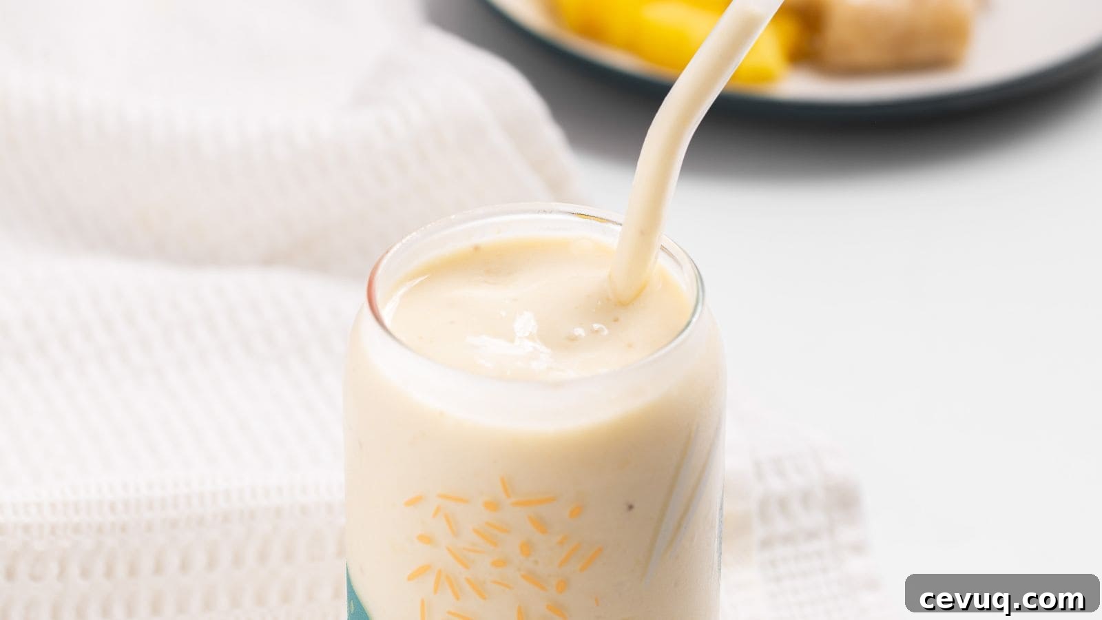 A close-up of a pineapple banana smoothie in a glass with a straw, surrounded by a white napkin with pineapple and banana in the background.