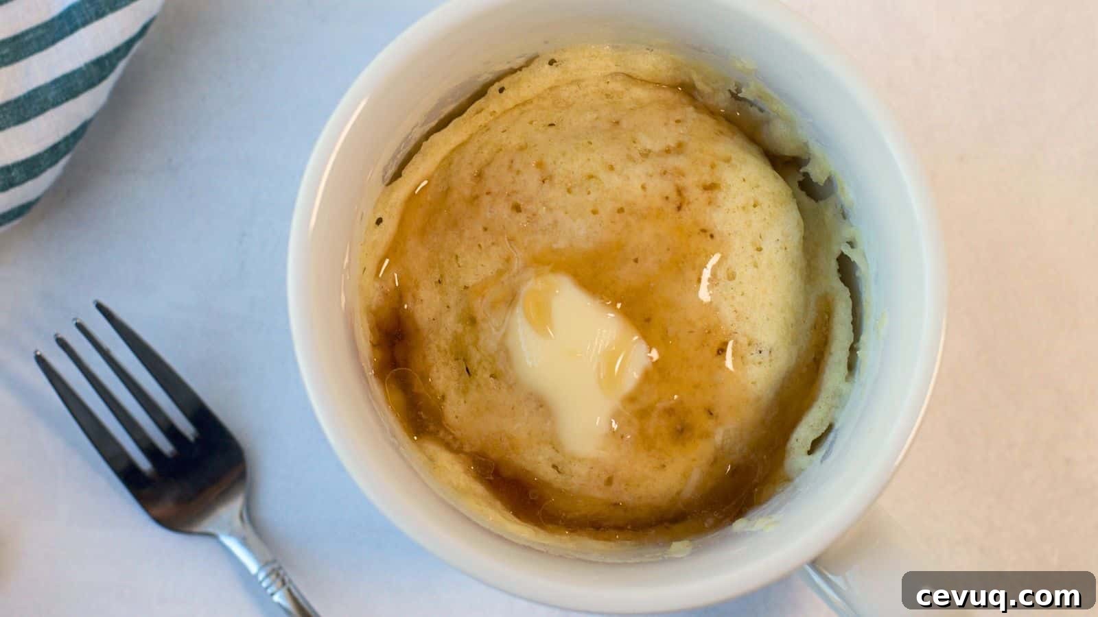 A pancake mug cake with syrup on top, viewed from above, next to a fork on a marble surface.