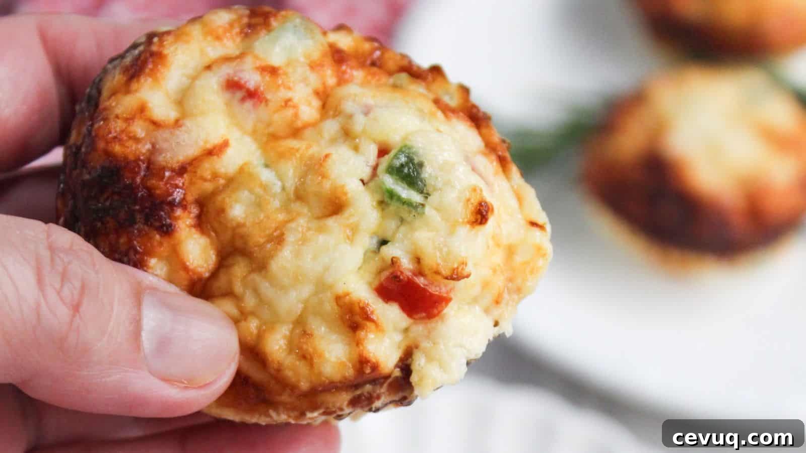 A close-up of a hand holding an egg white frittata muffin with visible toppings of cheese and chopped peppers.