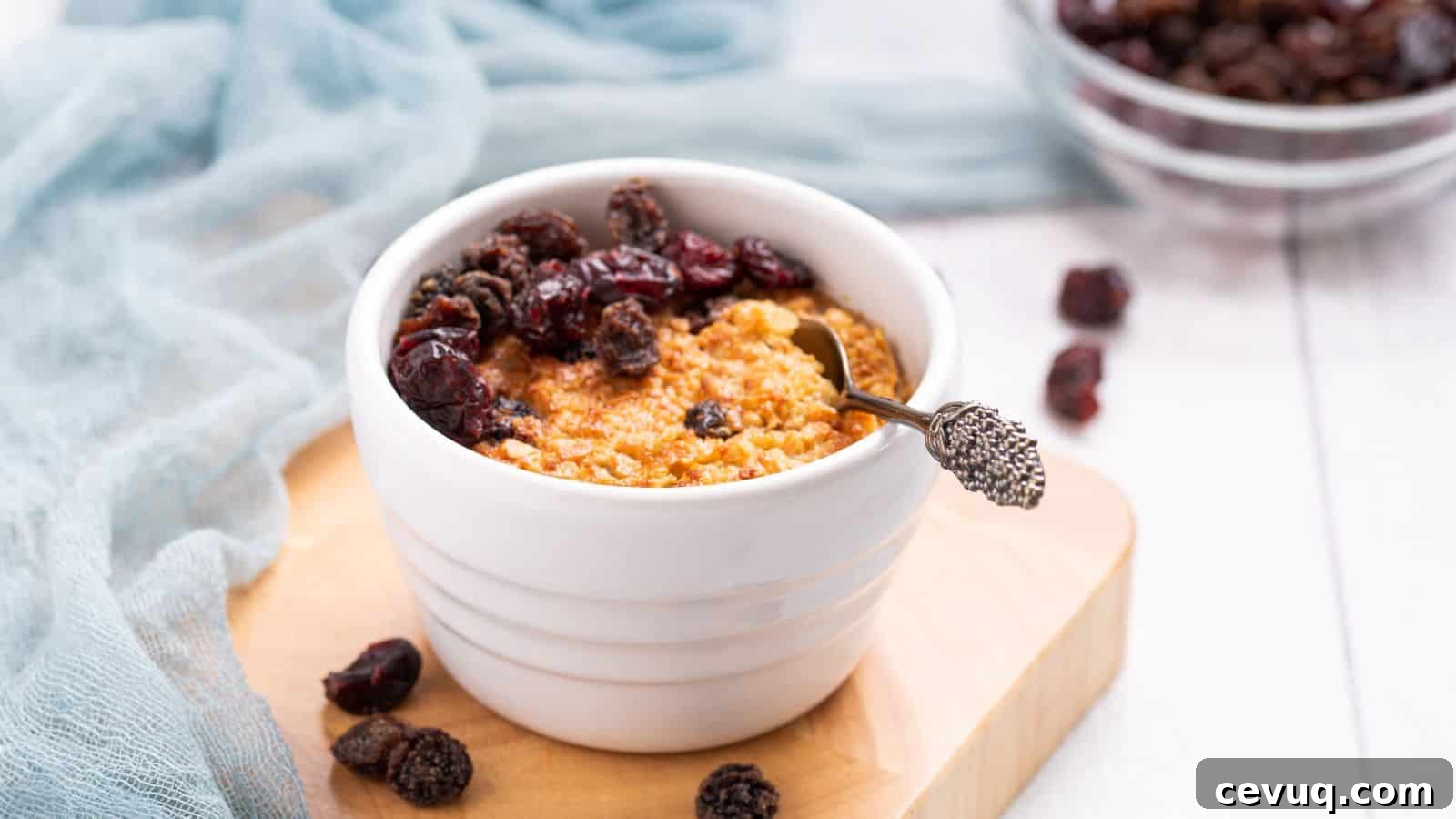 A bowl of baked oatmeal topped with raisins on a wooden table, accompanied by a blue cloth and spoon.