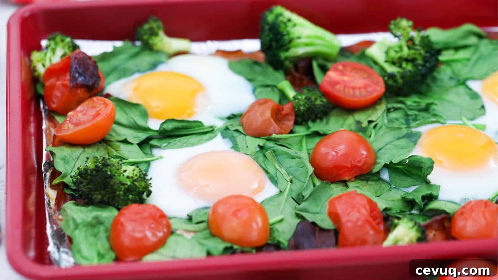 Baked eggs with spinach, broccoli, and cherry tomatoes in a red baking dish.