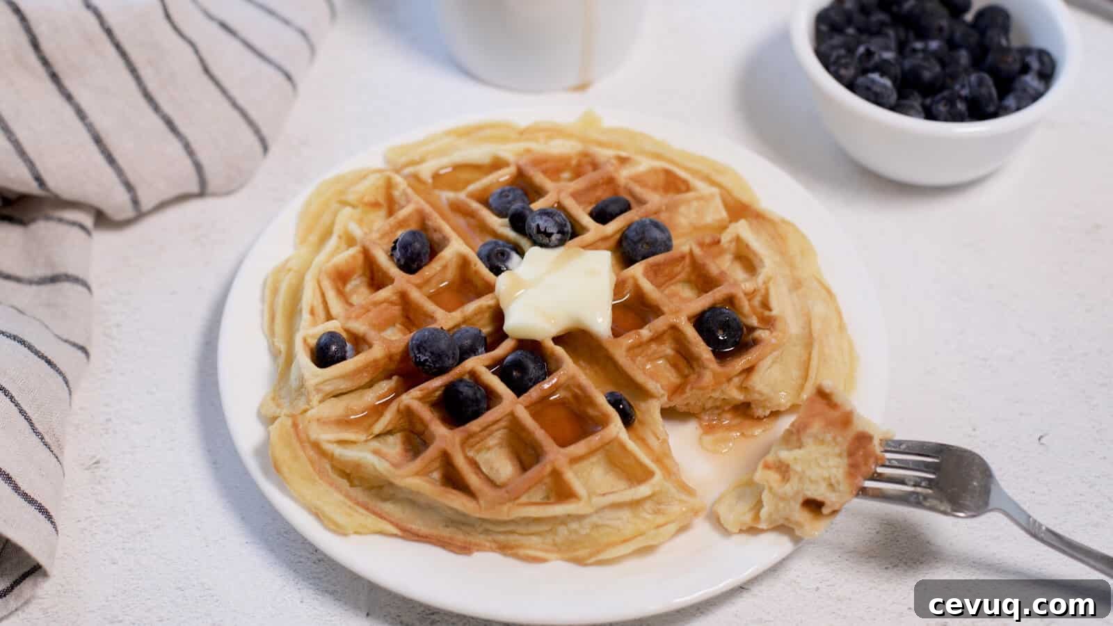 A freshly baked waffle topped with butter and syrup, garnished with blueberries, served with a side of more blueberries.