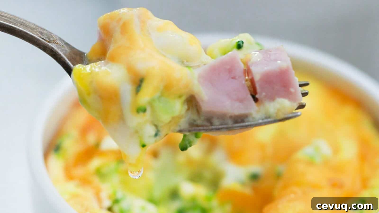 A fork holding a bite of mini frittata with ham and broccoli, with the dish in the background.