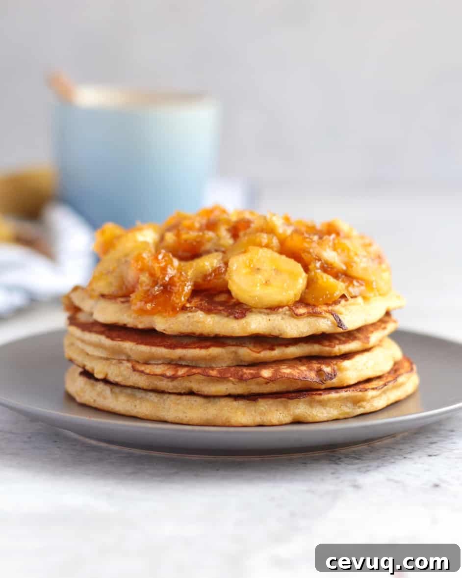 Stack of pancake topped with banana topping on a grey plate behind a white background.