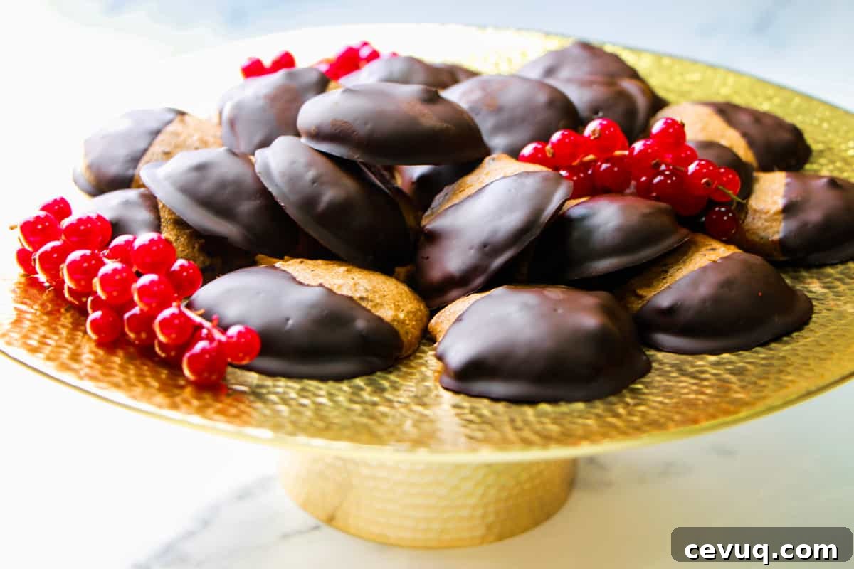 Photo of dark chocolate dipped almond cookies piled on a brass cake stand with redcurrants, all on a marble surface.
