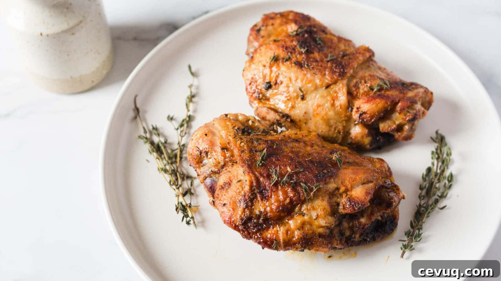 Two roasted chicken thighs on a white plate garnished with sprigs of thyme. A ceramic jar is partially visible in the background on the left.