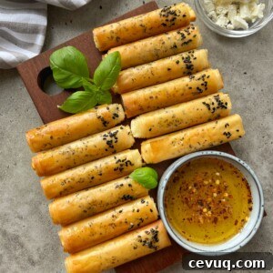 Golden, crispy Phyllo Feta Fries rolled and topped with black sesame seeds are arranged on a wooden board with basil leaves. A small bowl of seasoned olive oil and a bowl of crumbled cheese are nearby.
