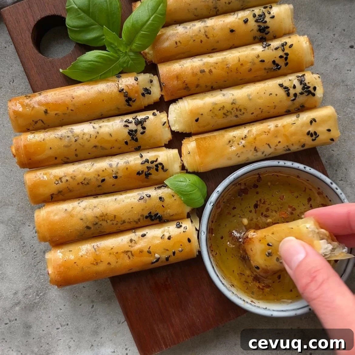 Crispy Phyllo Feta Fries topped with black sesame seeds are arranged on a wooden board with basil leaves, next to a bowl of hot honey.