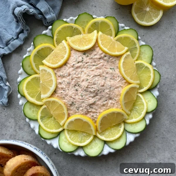 A round plate of creamy Hot Smoked Salmon Dip is garnished with alternating slices of lemon and cucumber in a circular pattern. A blue napkin, lemon halves, and toasted bread slices are nearby on a gray surface.