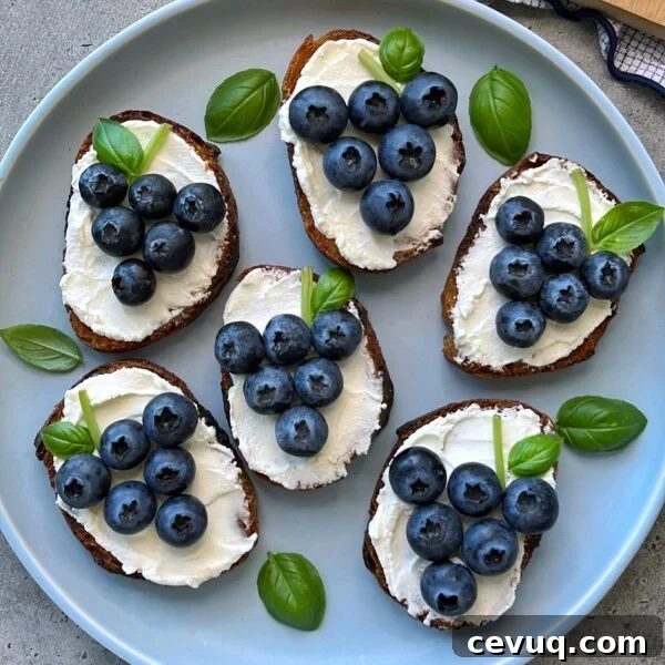 Six pieces of toast topped with goat cheese with honey and blueberries, arranged to resemble grape clusters, are garnished with fresh basil leaves on a light blue plate.
