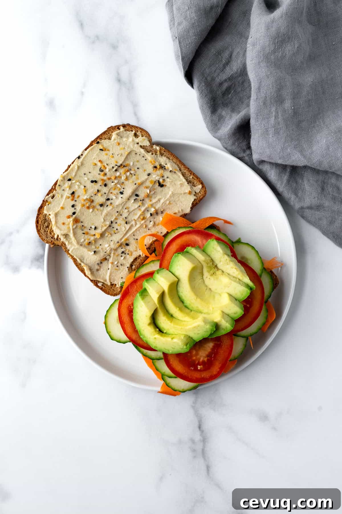 Open faced Cucumber Hummus Sandwich topped with carrots, tomato, and avocado on a white plate.