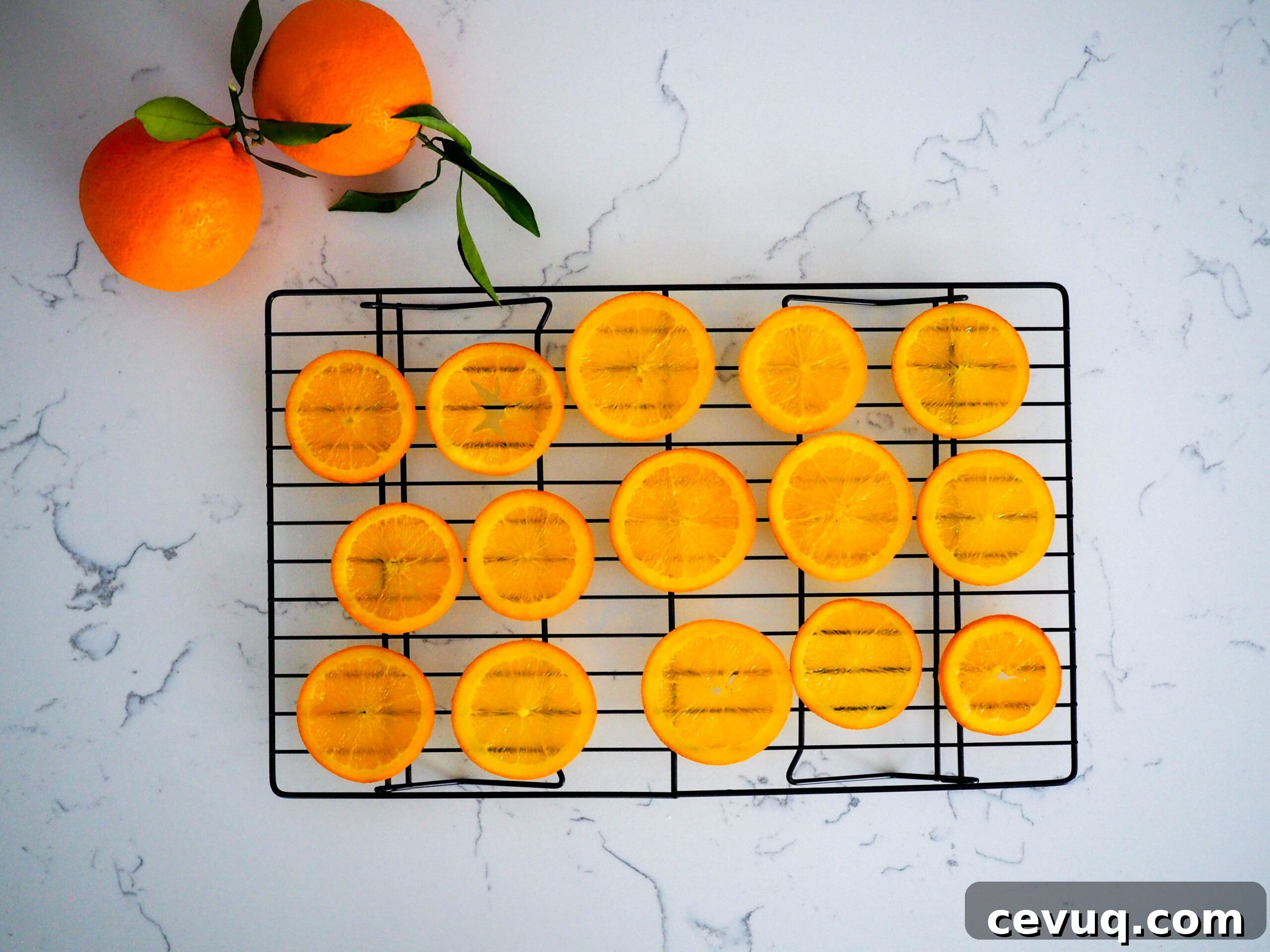 Fifteen orange slices dry on a wire rack with two leafy oranges beside them.