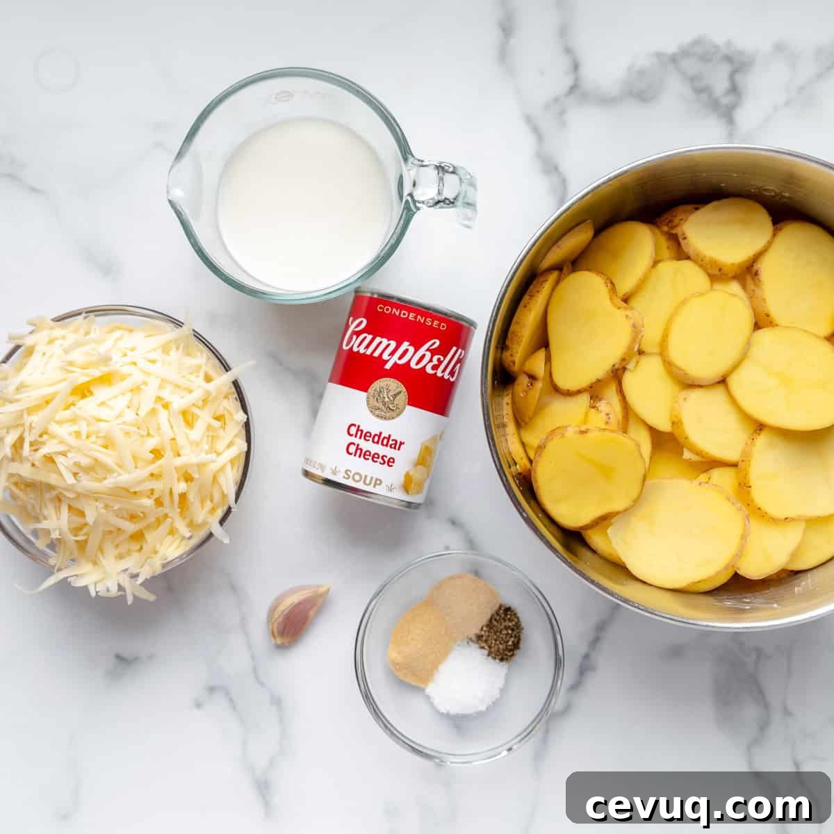 Sliced potatoes in a bowl, shredded cheese, a can of Campbell's Cheddar Cheese soup, milk, a clove of garlic, and seasoning on a marble surface.
