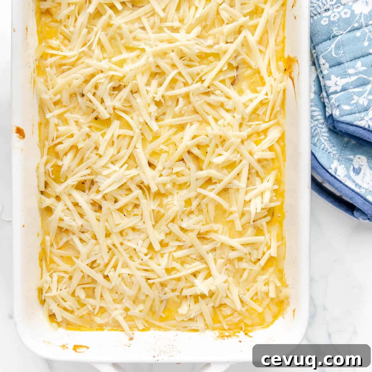 A casserole dish filled with shredded cheese on top of a layer of mashed potatoes, placed next to a blue patterned oven mitt.
