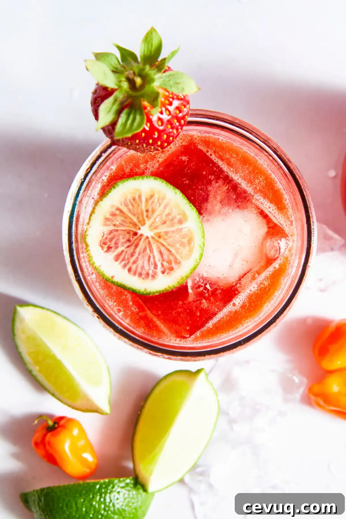 Overhead view of a spicy strawberry habanero margarita. The glass is garnished with a strawberry and surrounded by ice, habanero peppers and lime slices.