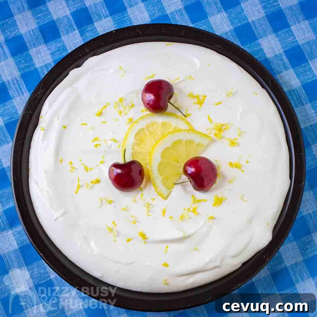 Overhead shot of a whole lemon pie on a black plate garnished with sliced lemons and cherries on a blue picnic cloth.