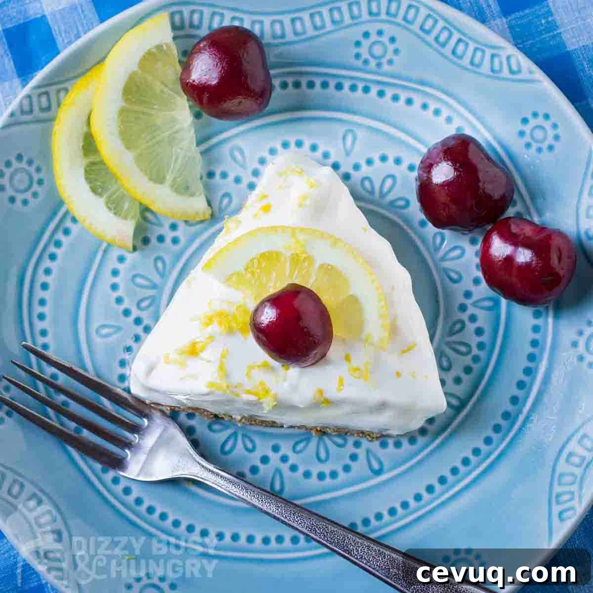 Overhead shot of a slice of lemon pie garnished with a whole cherry on a blue plate with a fork, lemon slices, and more cherries on the side.