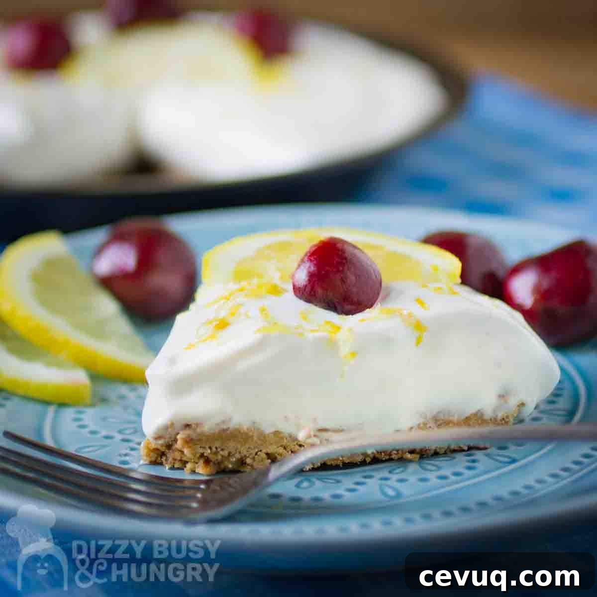 Side close up shot of a slice of lemon pie garnished with a whole cherry on a blue plate with a fork, lemon slices, and more cherries on the side.