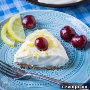 Close up shot of a slice of lemon pie garnished with a whole cherry on a blue plate with a fork, lemon slices, and more cherries on the side.