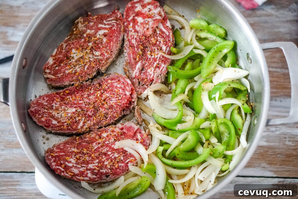Four steaks cook in a sauté pan with slices of green pepper and onion.