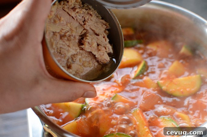 A can of tuna is added to a spicy stew in a pot.