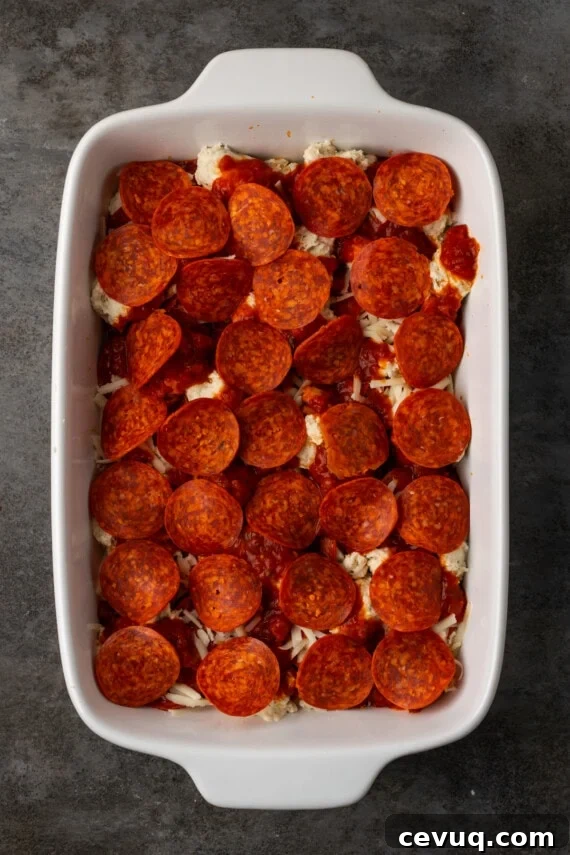 A final layer of pepperoni is added to pizza casserole in a baking dish.